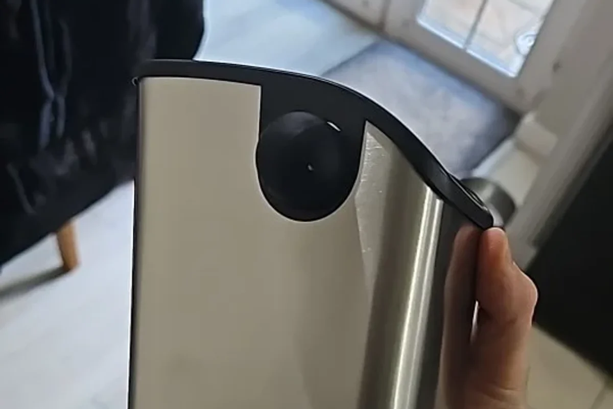 The brushed stainless steel body of the Barwright knock box held in one hand, showing the black collar and the round mount that carries the knock bar